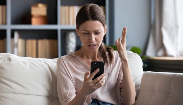Confused angry woman having problem with phone, sitting on couch at home, unhappy young female looking at screen, dissatisfied by discharged or broken smartphone, reading bad news in message